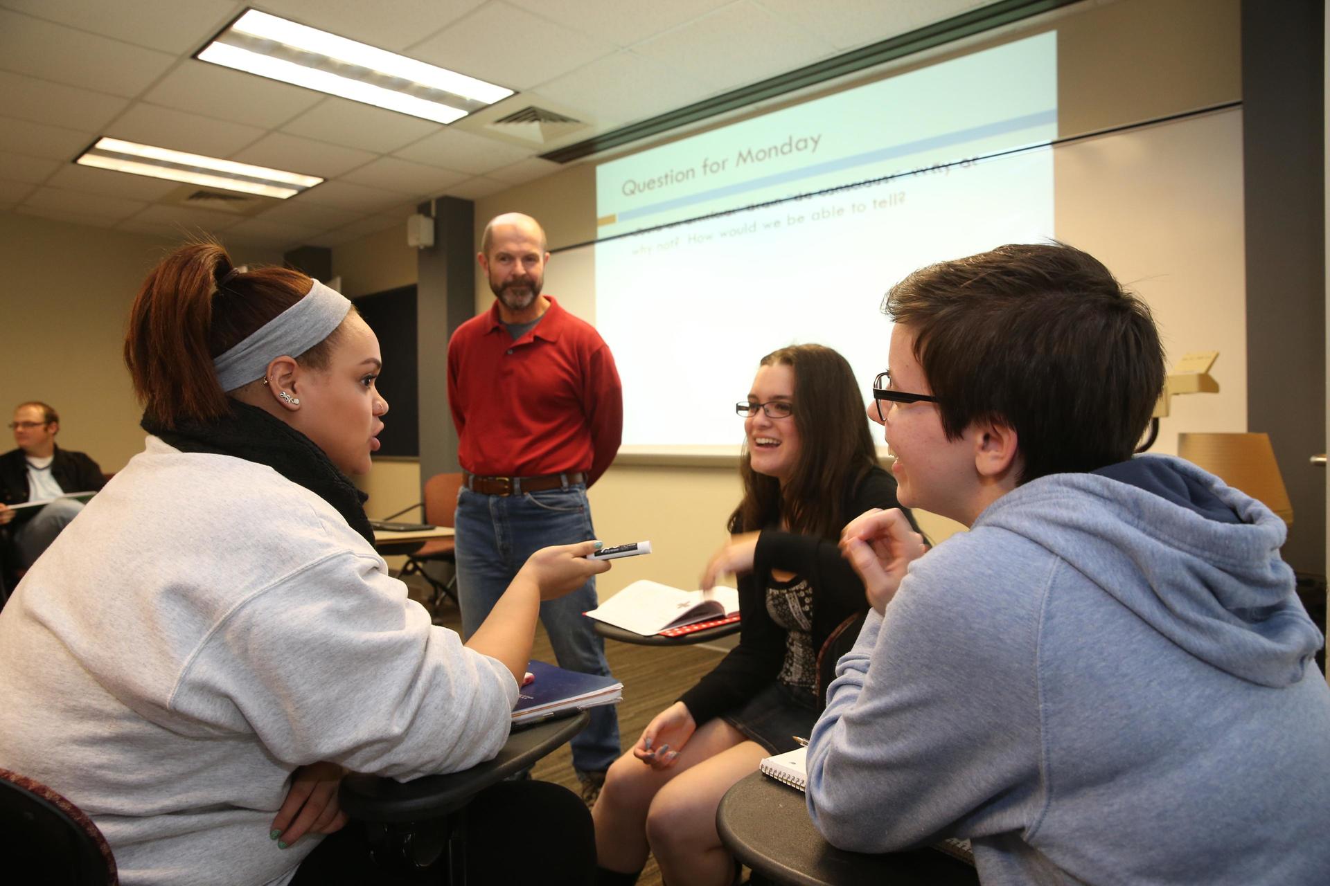 A teacher stands in front of three students in conversation.