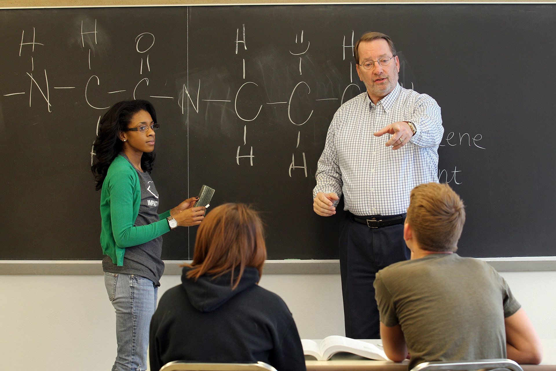 Two people stand in front of a chalkboard and two students.