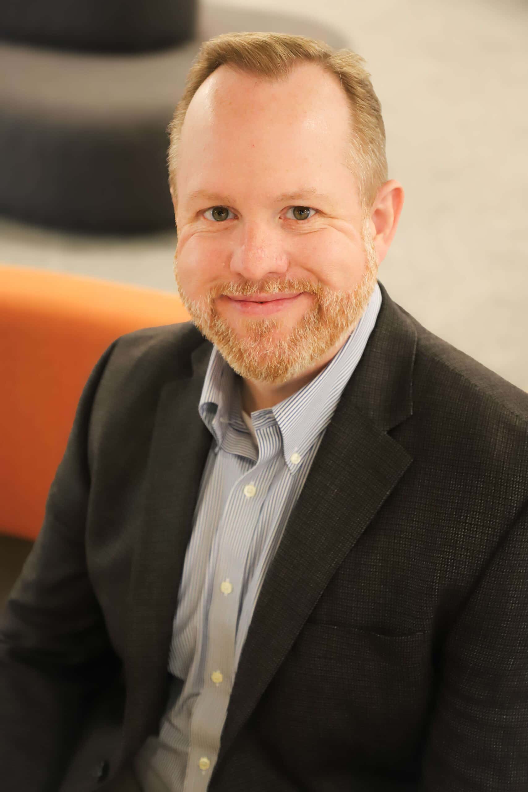 A bearded man in a dark gray sport coat and blue striped shirt, seated in an orange chair, with a friendly and engaged expression. 