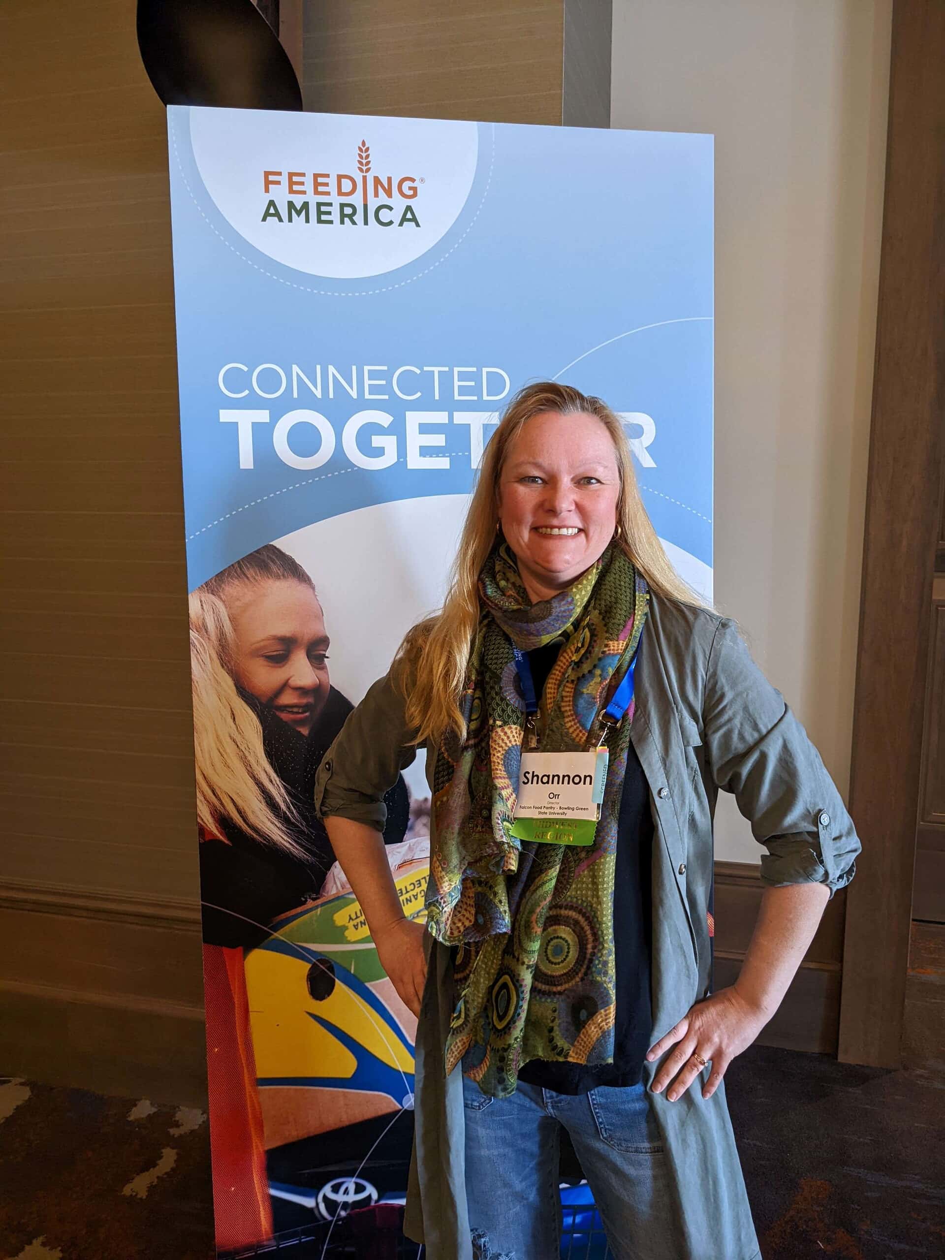 A smiling woman wearing a patterned scarf and long button-down shirt, standing in front of a poster for Feeding America.