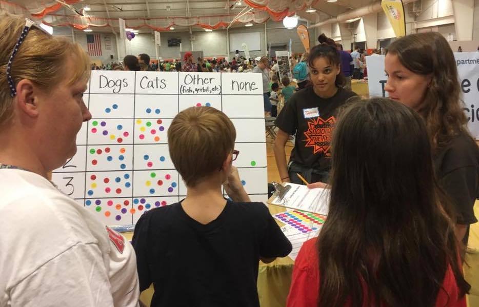 Two undergrad students speak with three STEM participants -- 1 adult, frame left, and two children, frame center