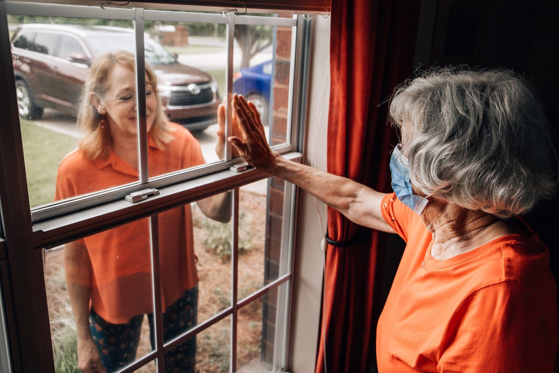 A baby boomer daughter visiting her elderly shut-in mother in her 80's during quarantine from COVID-19 coronavirus through a window as to not catch this contagious disease