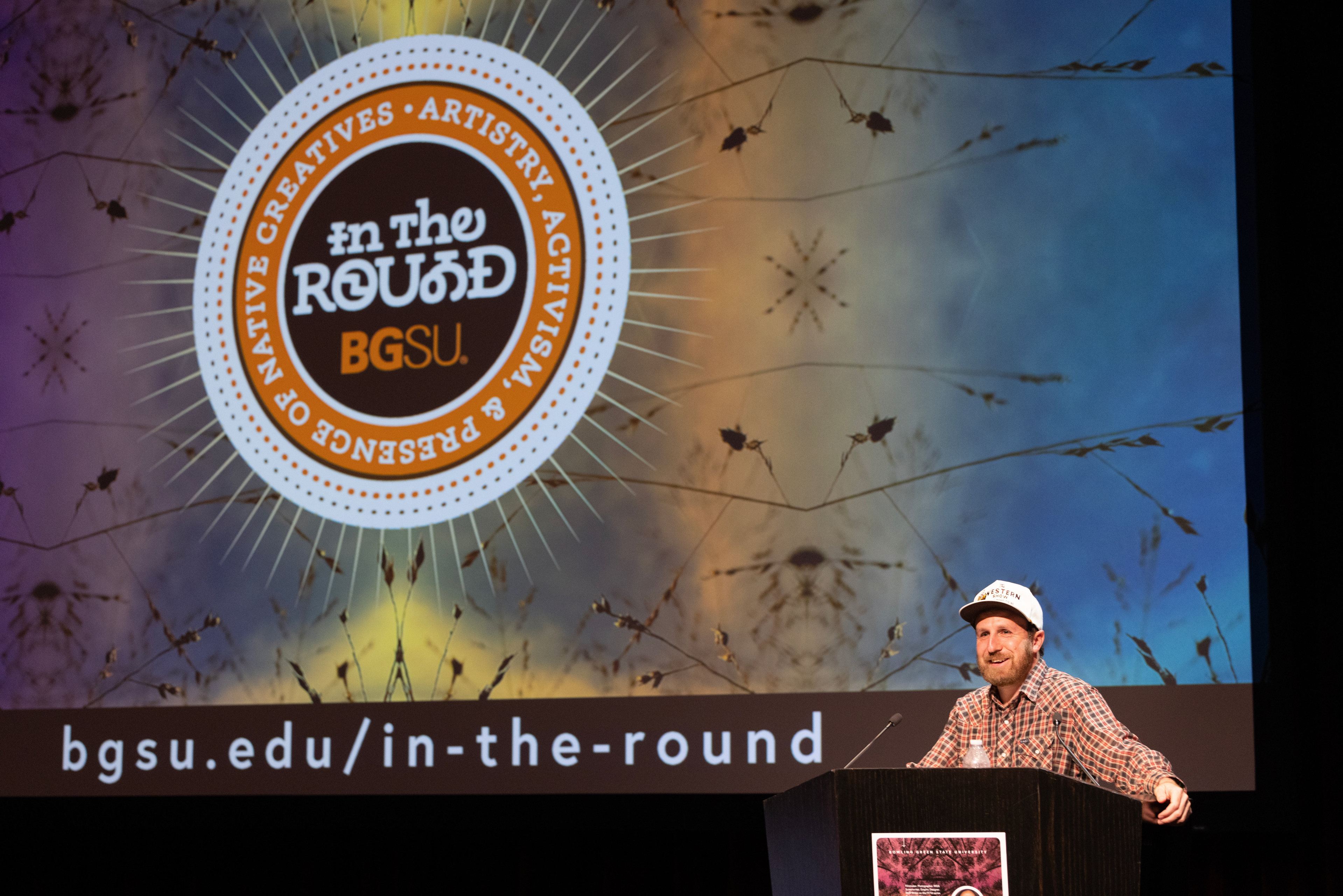 Ryan RedCorn smiles in front of a screen with the BGSU In The Round logo.