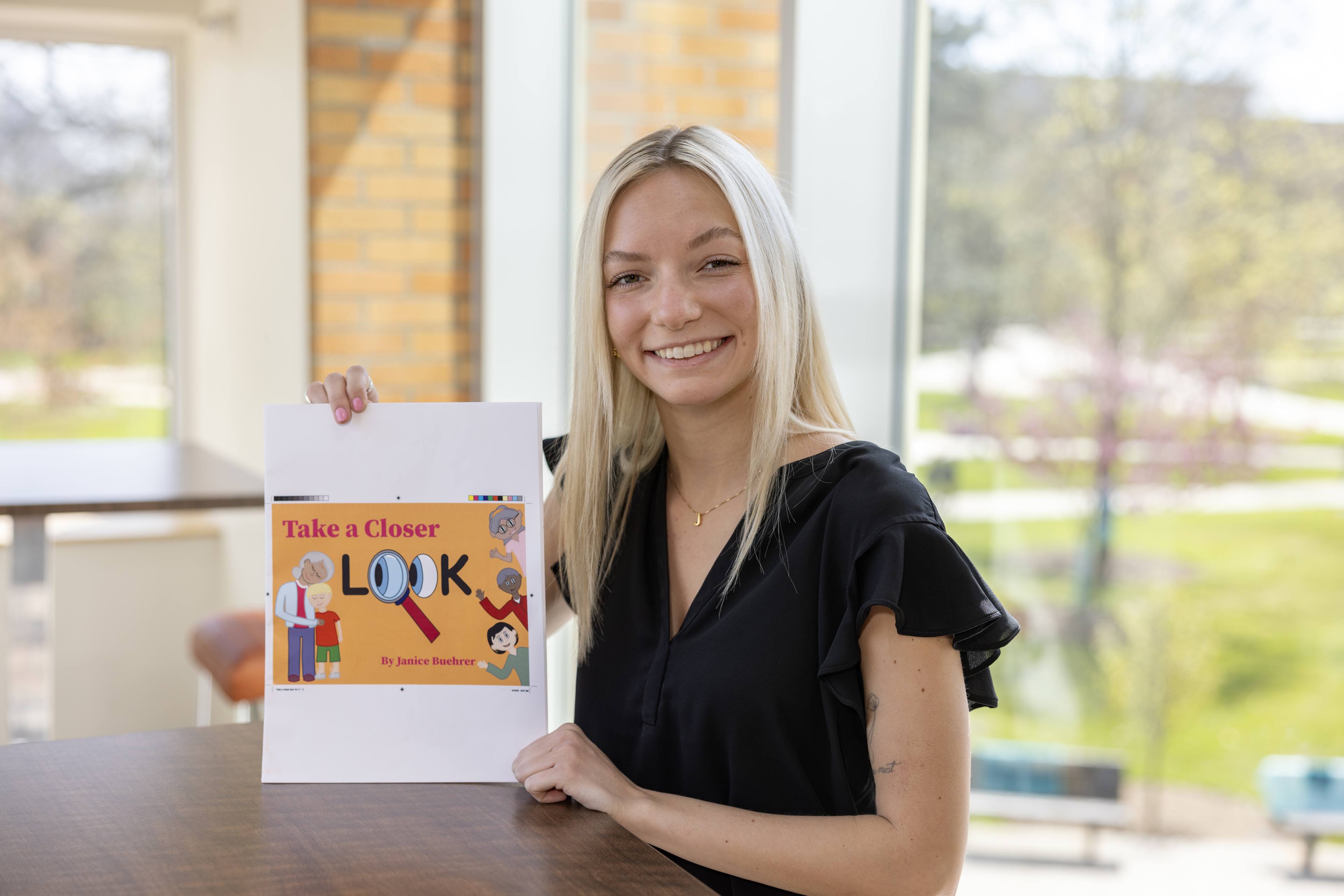 A female student sits at a table and holds up her book, titled "Take A Closer Look" 