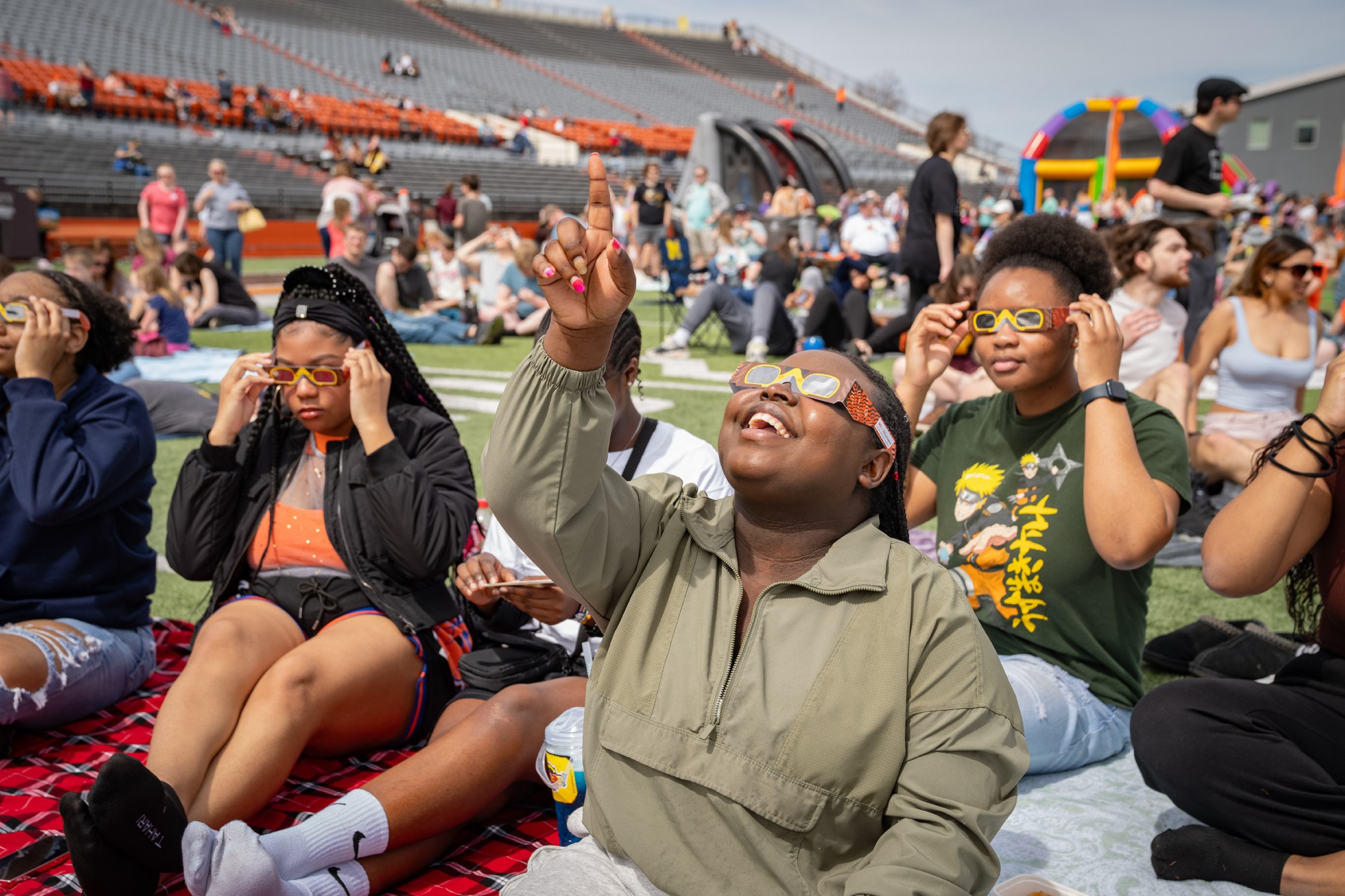 A group of students sit on a blanket and wear eclipse glasses 
