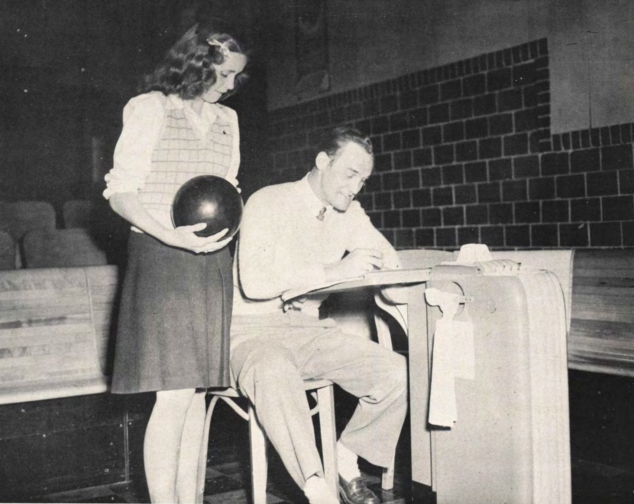 A young woman holds a bowling ball while a young man sits at a seat on the alley