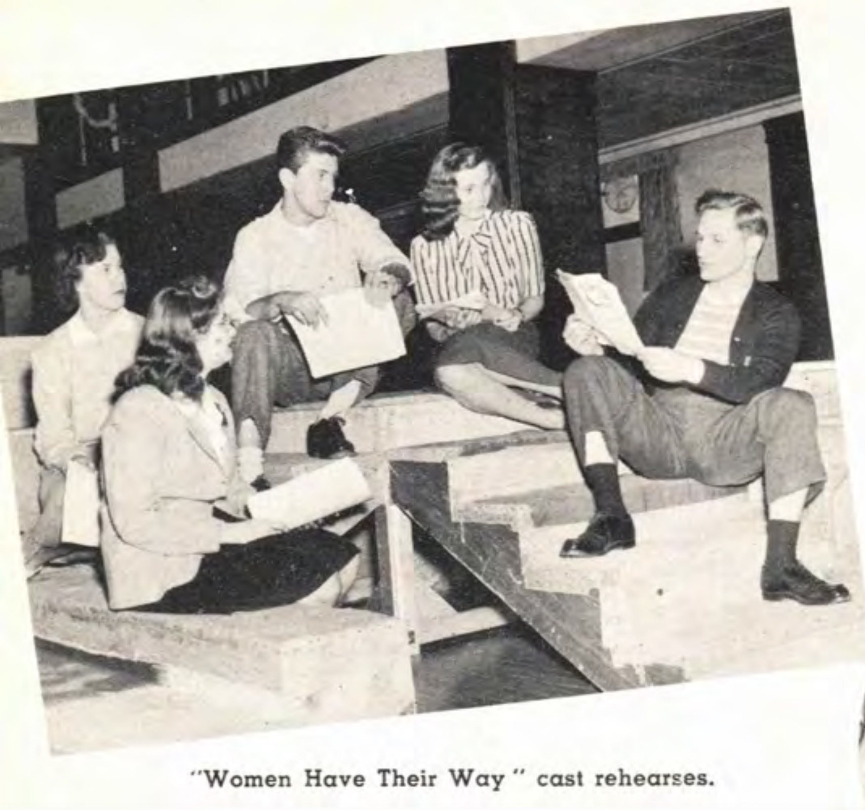 A group of five young people sit on steps and read scripts