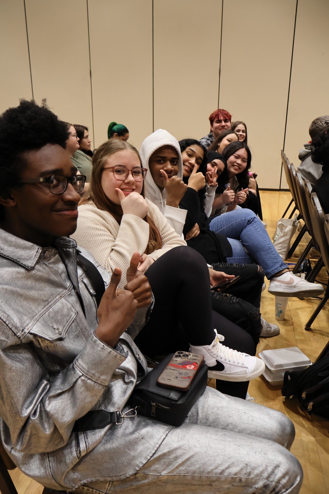 A row of students seated in folding chairs give thumbs up