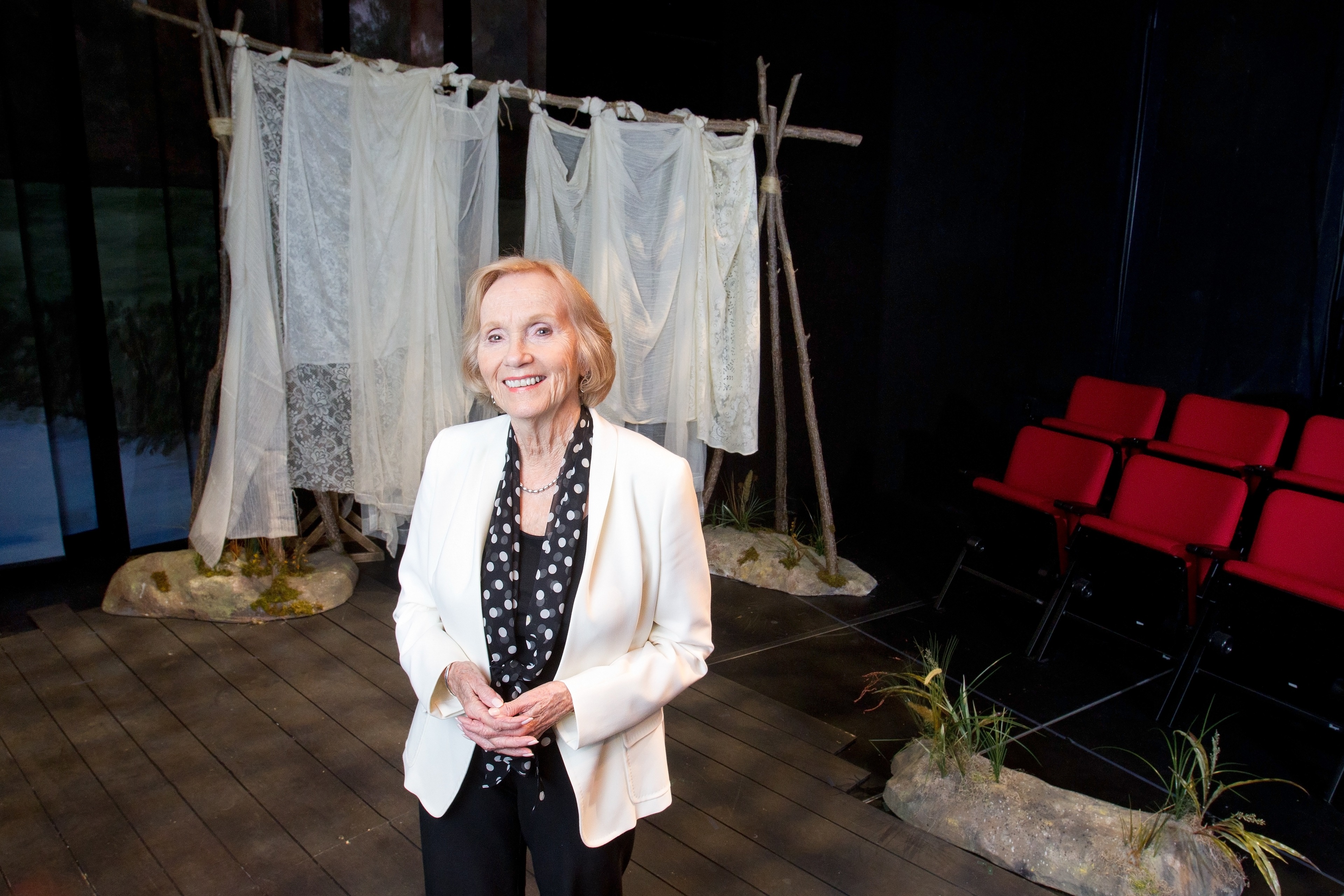  A woman in a white blazer and black with white polka dots scarf poses for a photo in a theater