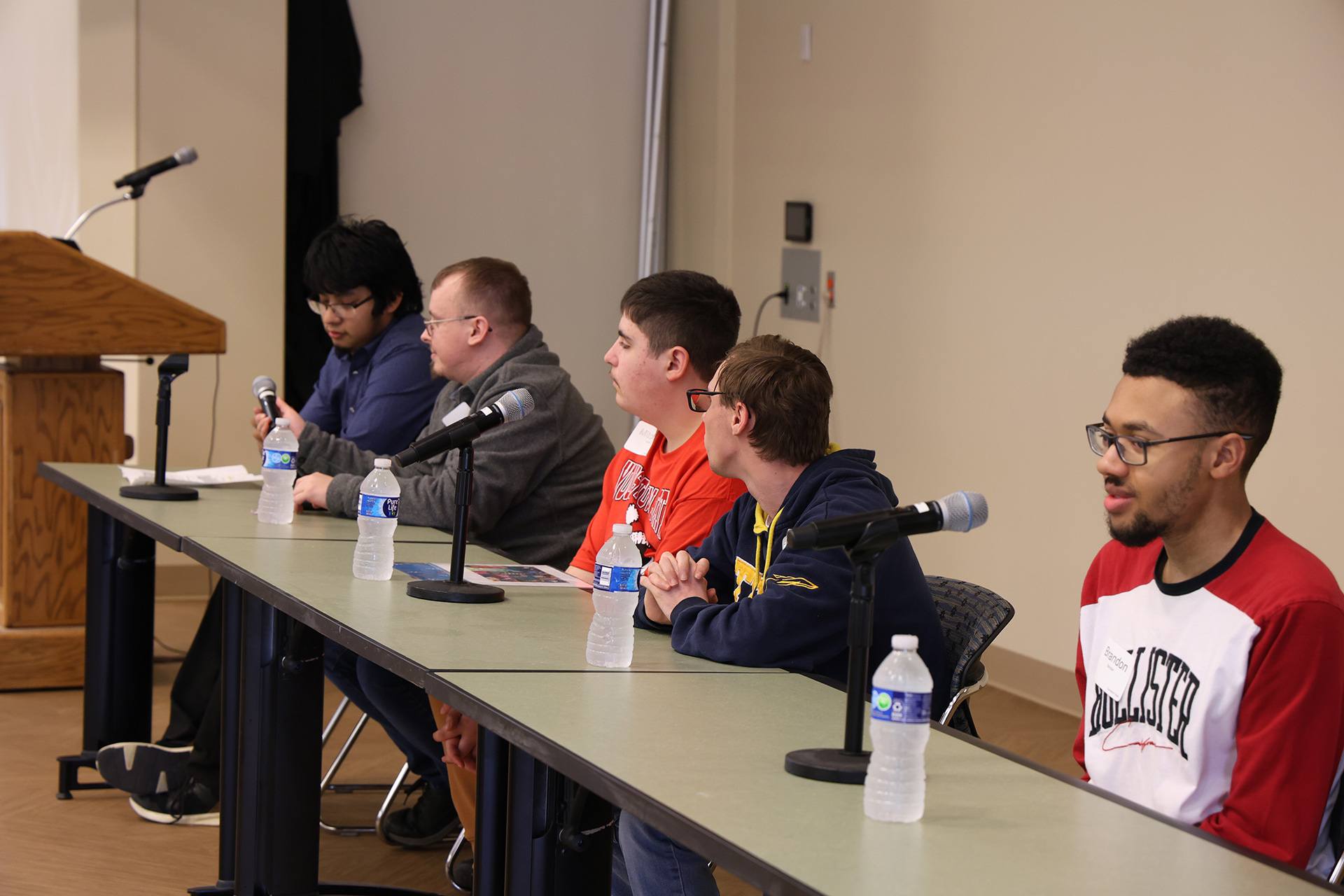 A group of students sit at a panel of tables with microphones
