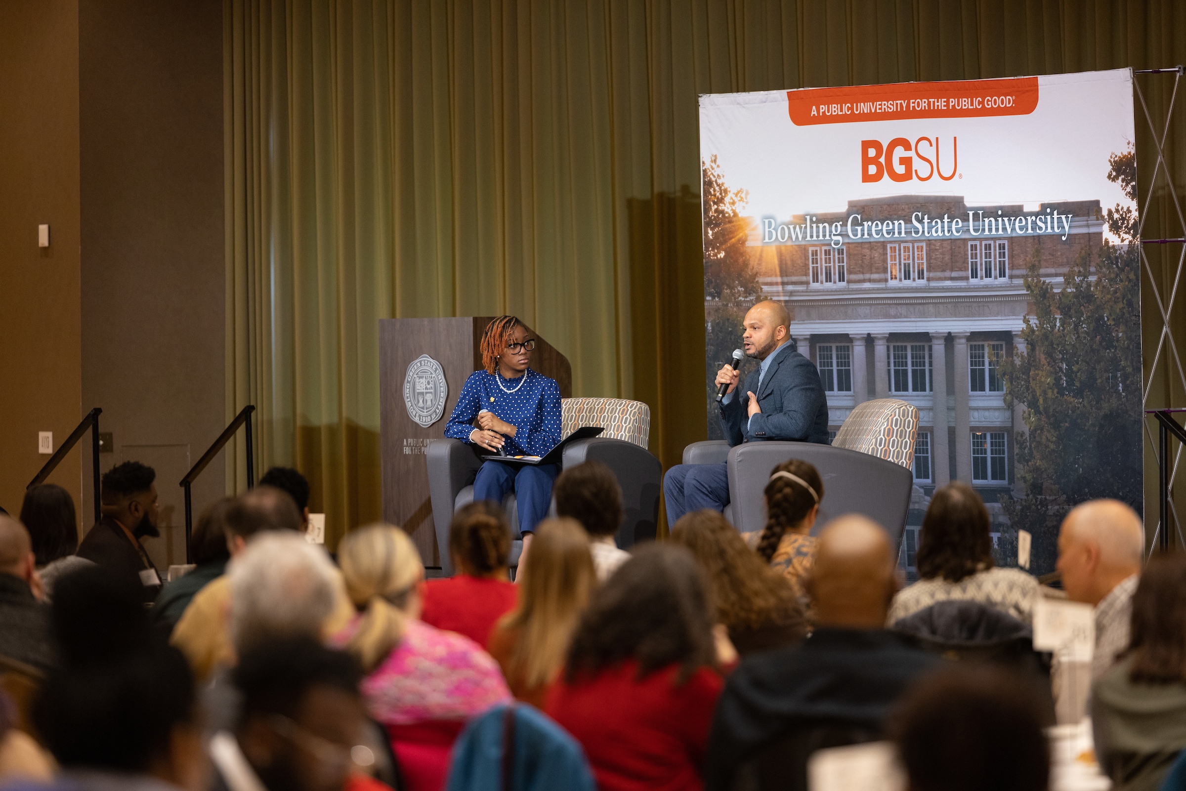 A man and woman sit on a stage talking to a crowd of people