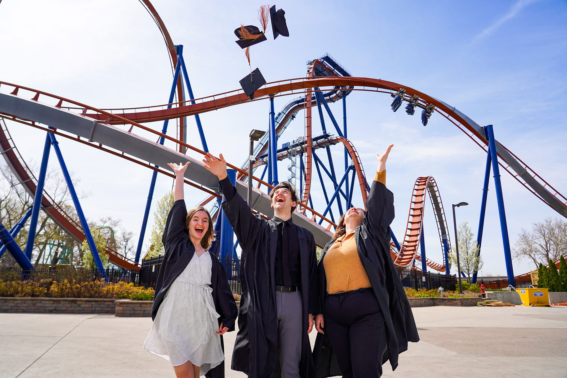 Three college graduates throw their caps in the air with roller coasters in the background