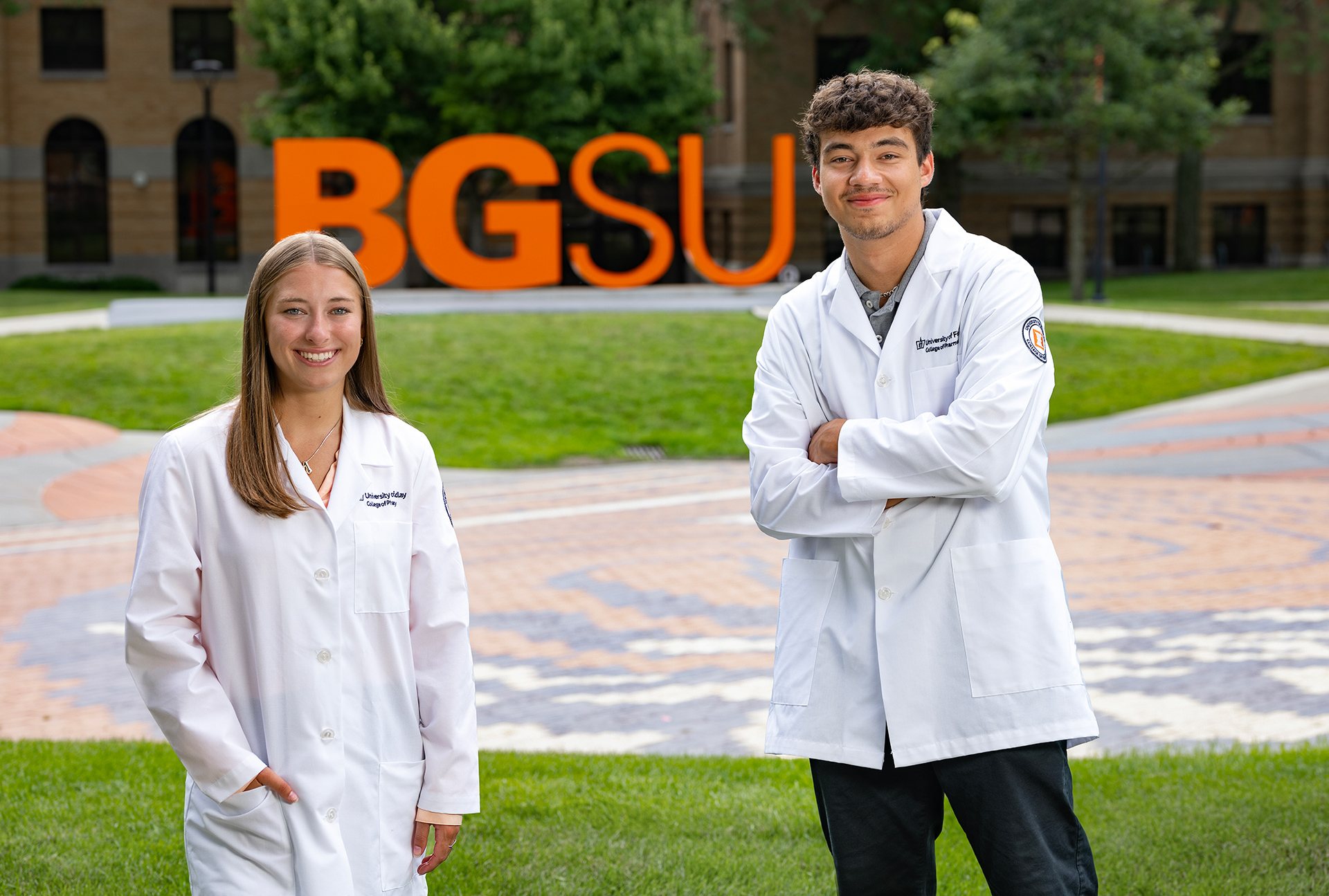 Two people stand in white medical coats in front of "BGSU" letters