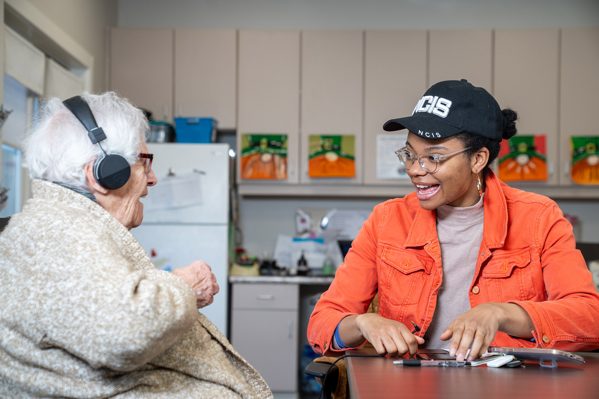 Two people interact at a table while one listens to music wearing headphones