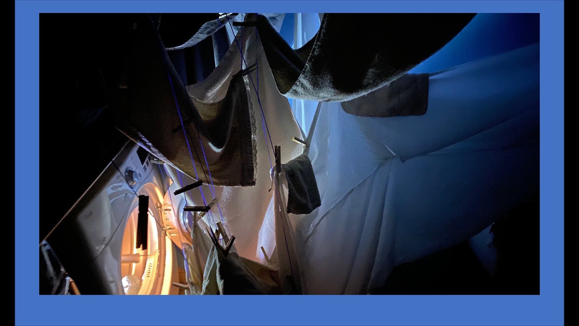 dark eerie interior of laundry room with handing cloth crowding toward blue light coming from inside or washing machine