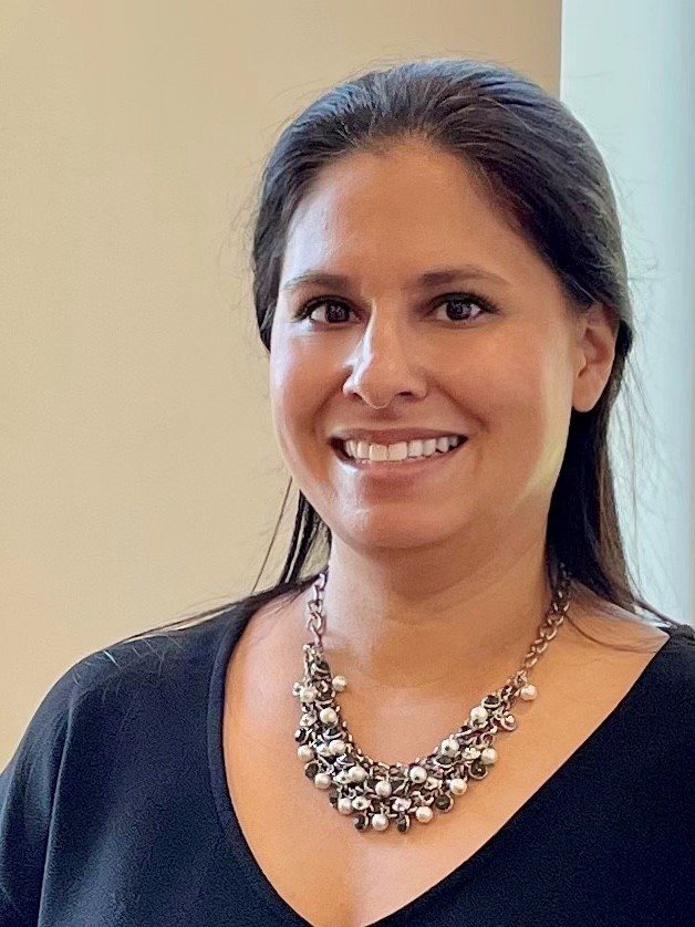 Picture of Marisa Cline. She has her long dark haired pulled back away from her face. She is wearing a large decorative necklace and dark colored shirt. She is also standing against a light colored wall.