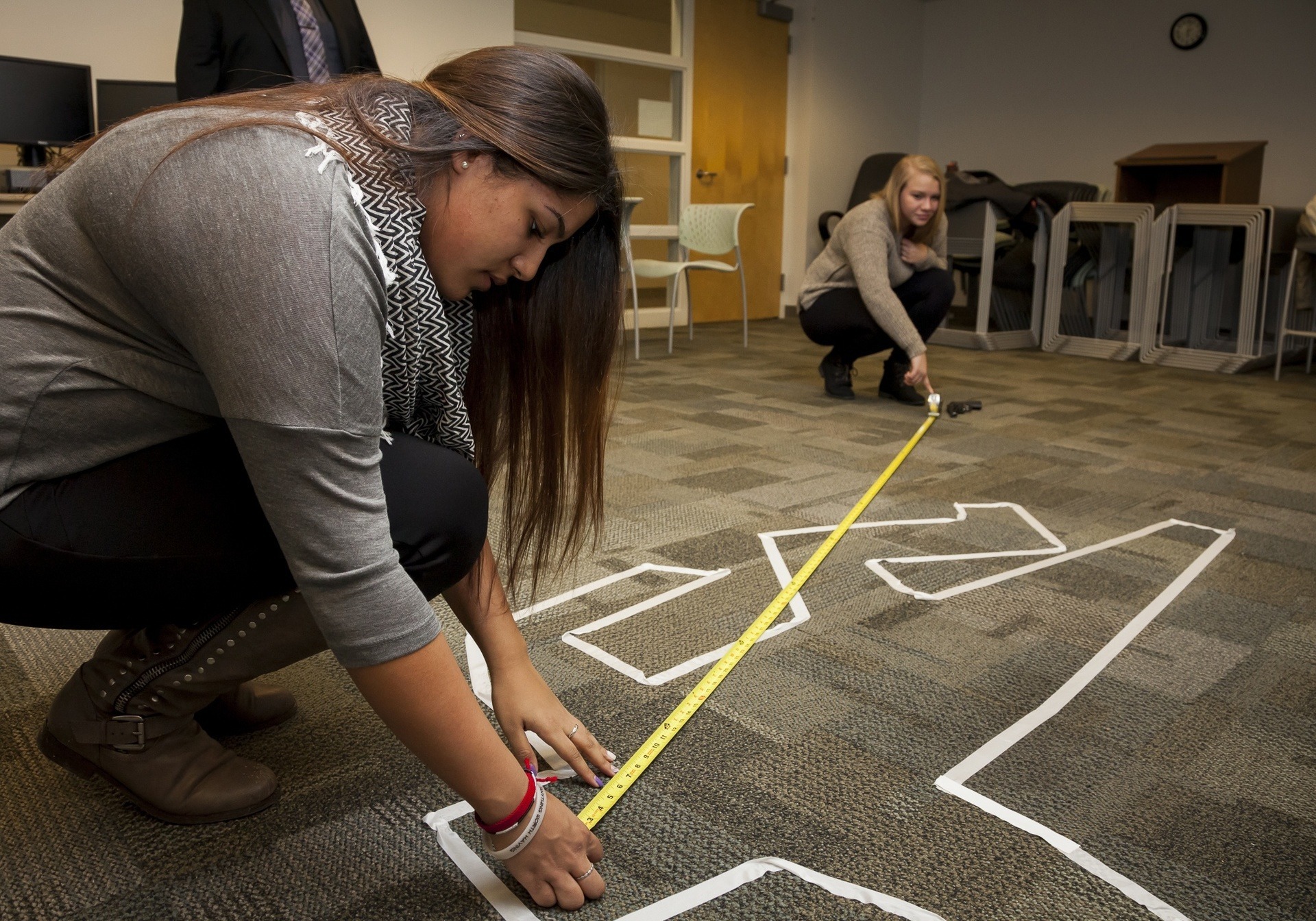Two female BGSU forensic investigation students measure the distance from a body to a firearm in an on-campus crime scene lab.