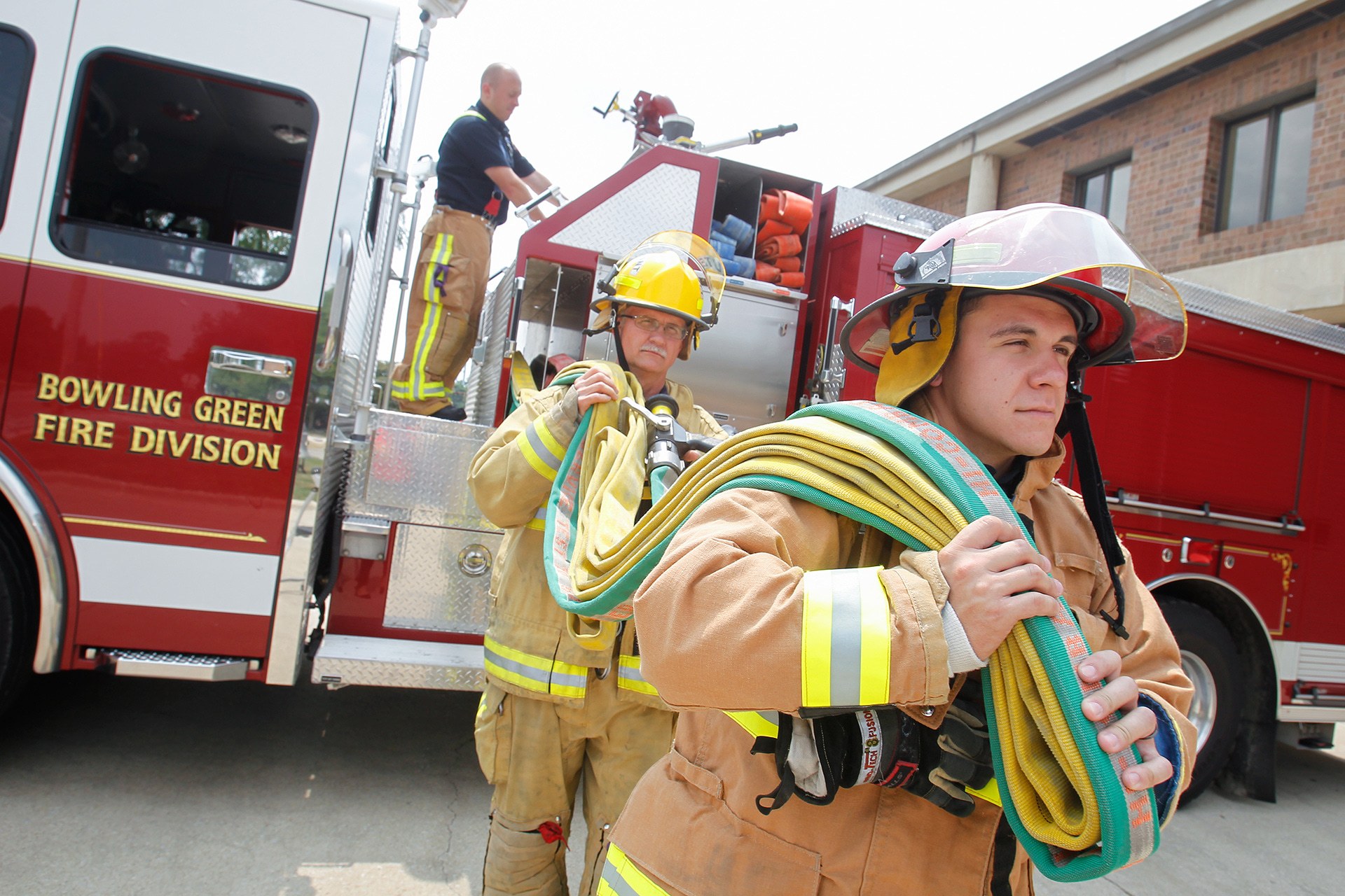 3 firefighters pull a hose away from a firetruck.